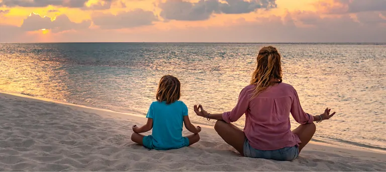 A mother and child meditate peacefully on a beach during sunset, embodying the tranquility of Suniai Oliva.