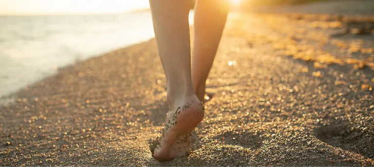 A close-up view of bare feet walking on a sandy beach during sunset. The sunlight casts a warm glow on the scene, and the sand shows slight footprints in the foreground. The ocean can be seen in the background, with gentle waves lapping at the shore.