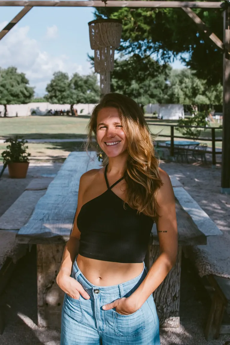 Aina, the Kitchen & Booking Manager at Suniai Oliva, smiles warmly in a sunny outdoor setting by a rustic wooden table.
