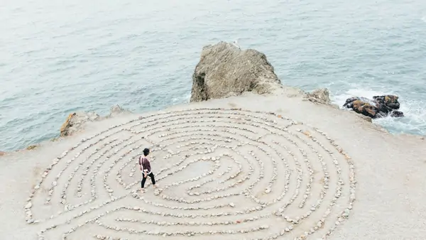 A person walks through a stone labyrinth by the ocean on a rocky shore under a cloudy sky.