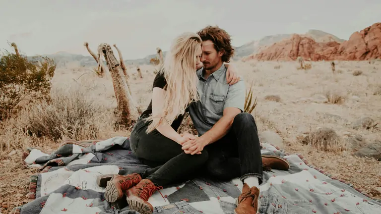 A couple sits on a blanket in a desert landscape, sharing an intimate moment amid cacti and rocky formations.
