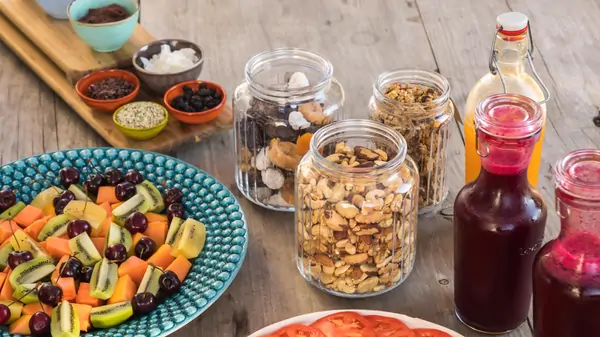 A colorful spread featuring a fruit salad, granola in jars, assorted sauces, and sliced tomatoes on a wooden table.
