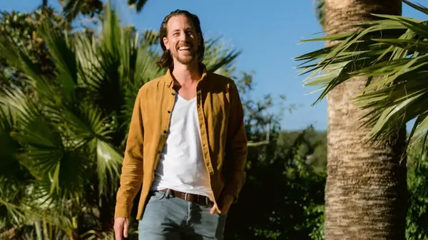 A smiling man stands near a tall palm tree, surrounded by lush greenery under a clear blue sky.