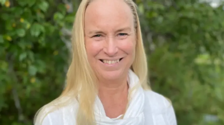 A smiling woman with long blonde hair stands outdoors in front of greenery, wearing a white top and scarf.