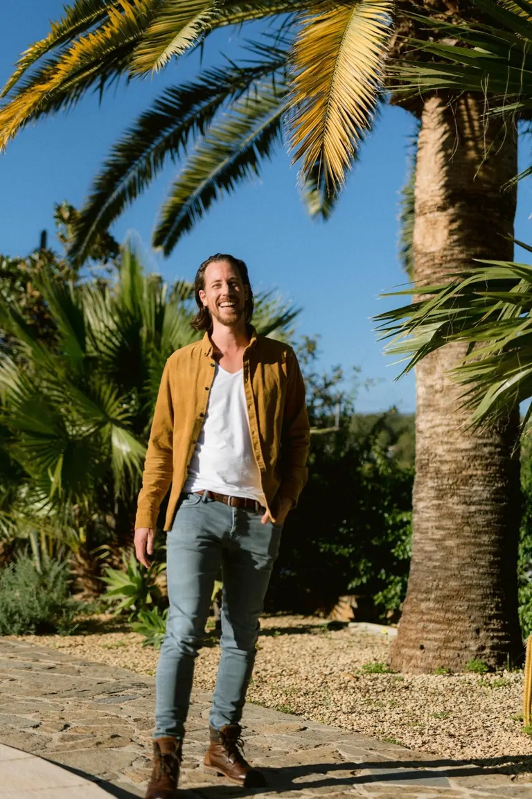 A smiling man stands near a tall palm tree, surrounded by lush greenery under a clear blue sky.