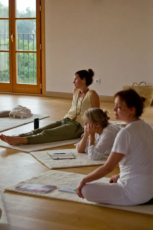 Group of women sitting on mats in a serene yoga studio, engaged in meditation or mindfulness practice. Natural light filters in.