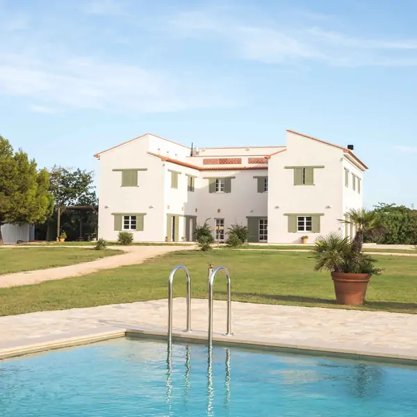 A modern white house with green shutters, set in a grassy field beside a swimming pool under a clear blue sky.